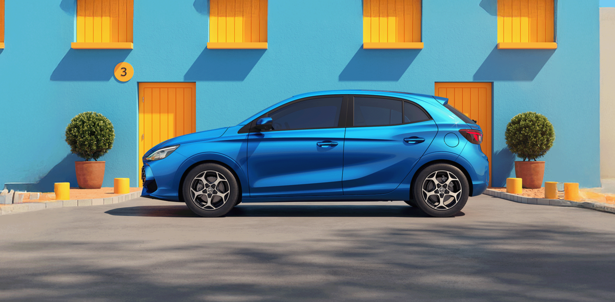 MG3 in Como Blue, seitlich stehend vor hellblauem Gebäude mit gelben Fenstern und Türen